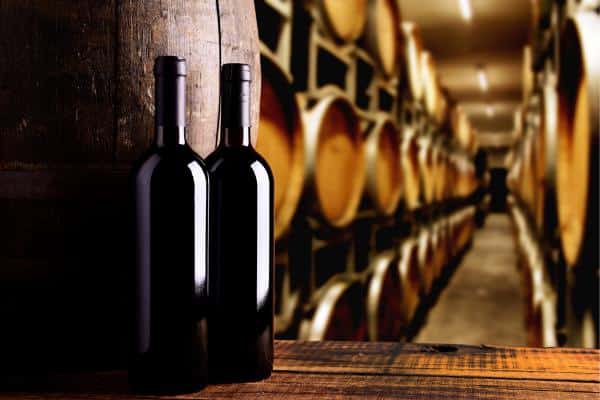 Dos botellas de vino apoyadas en una mesa, en una bodega llena de barricas.